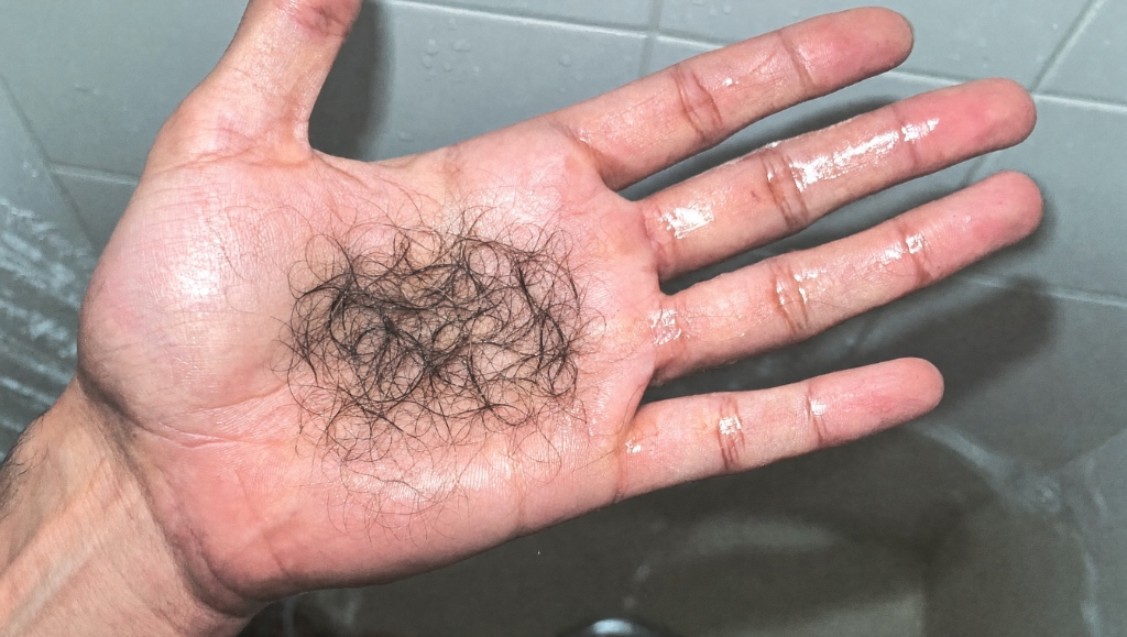 A bunch of shed hair resting in the palm of a wet hand in front of bathroom tiles.